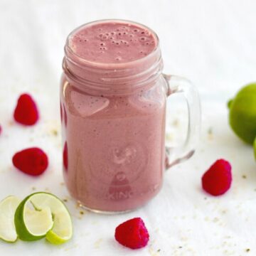 pink smoothie in glass jar with handle surrounded by random raspberries and parts of lime