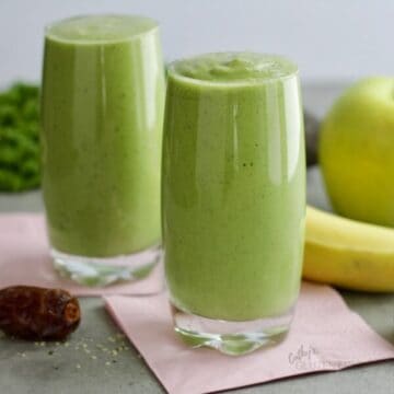 two glasses of green smoothie on pink napkins with a date, banana, and green apple to the side