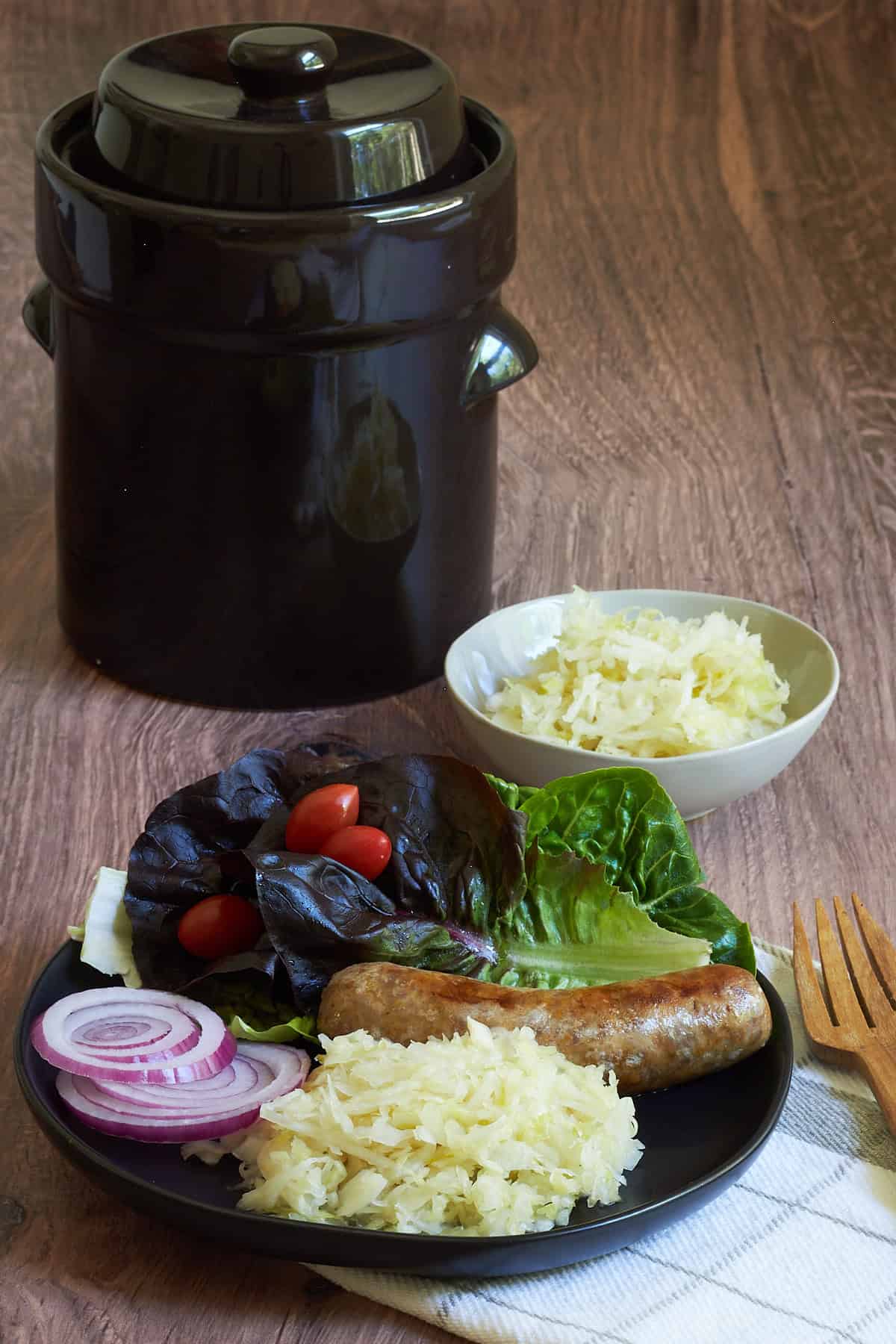 raw sauerkraut on black plate with sausage and salad, fermentation crock in background.