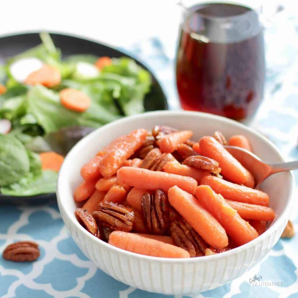 Maple Glazed Carrots with Pecans in white bowl with salad and syrup