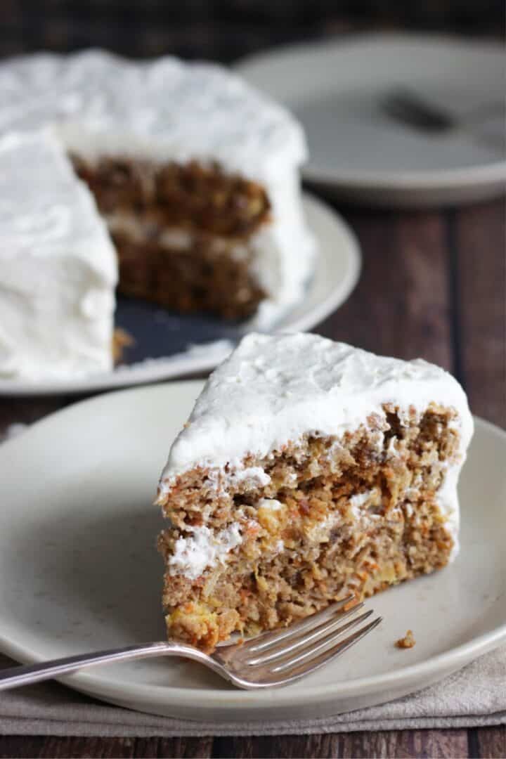 wedge of frosted carrot cake on plate with fork with remaining cake and extra plate and fork in background