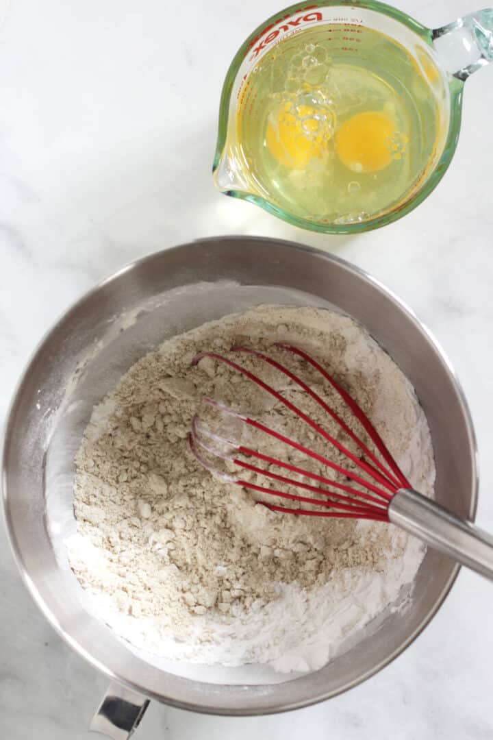 red whisk in dry flour mixture in mixing bowl with eggs in cup to side.