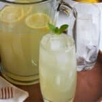 freshly squeezed lemonade on ice in glass garnished with mint in front of pitcher.