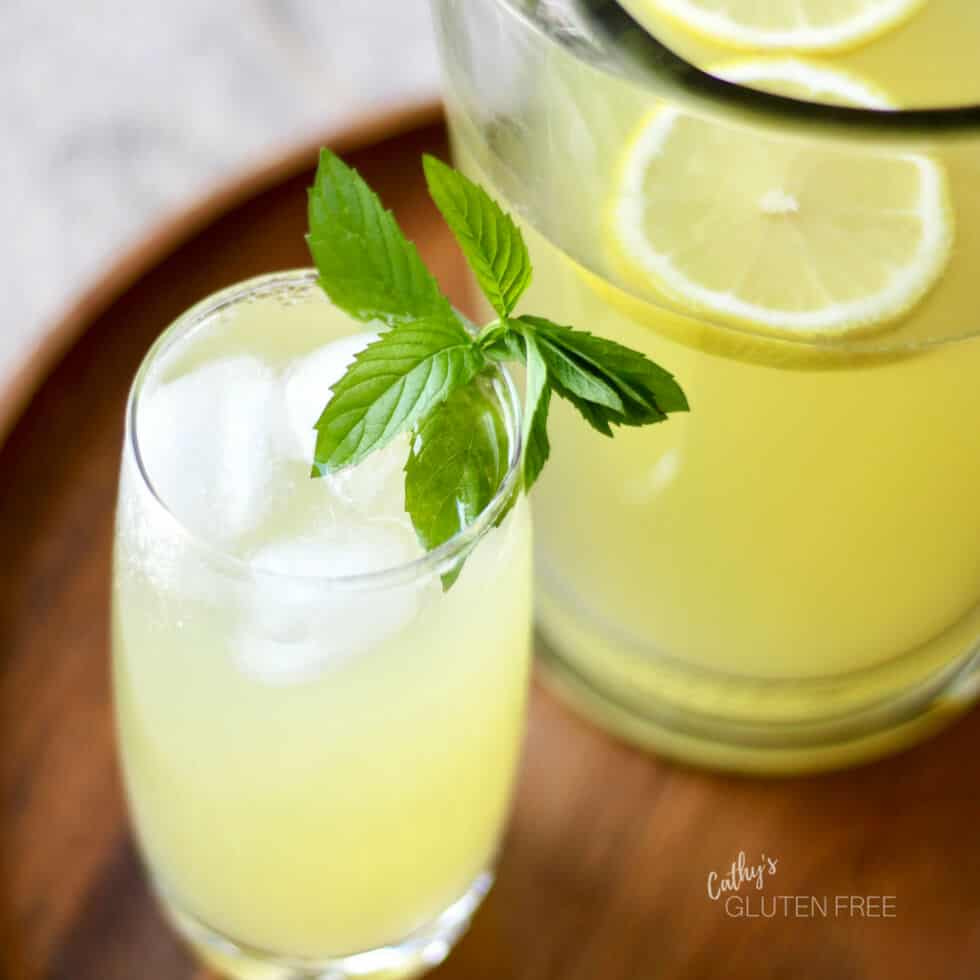 a glass of lemonade with ice and a sprig of mint sit on a wooden tray by a pitcher of lemonade