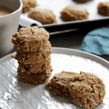 ginger cookies stacked on a white plate with cup of coffee and more cookies on baking tray in background