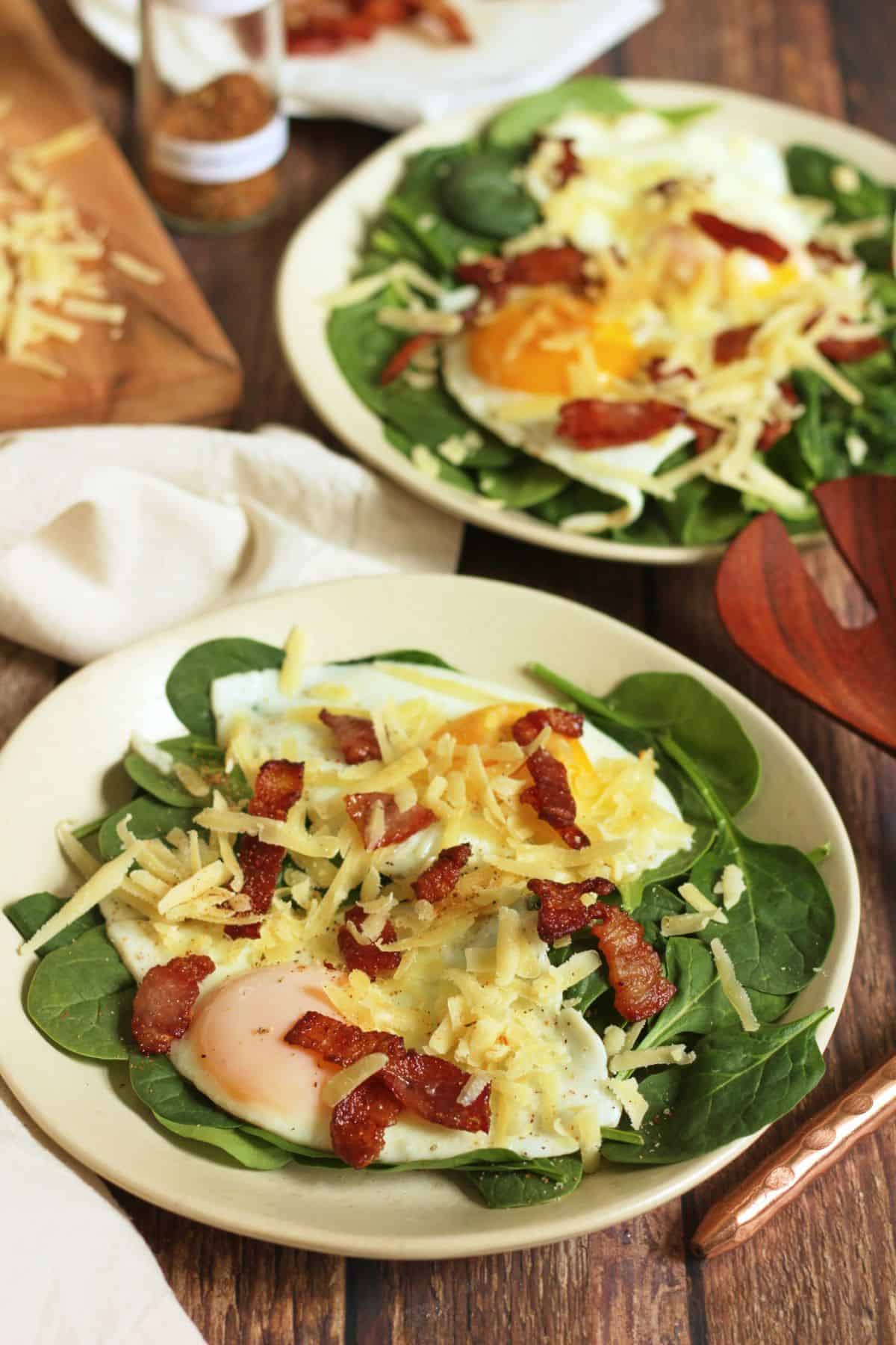 two plates of salad with chopped bacon, shredded cheese, and fried eggs layered over fresh spinach.