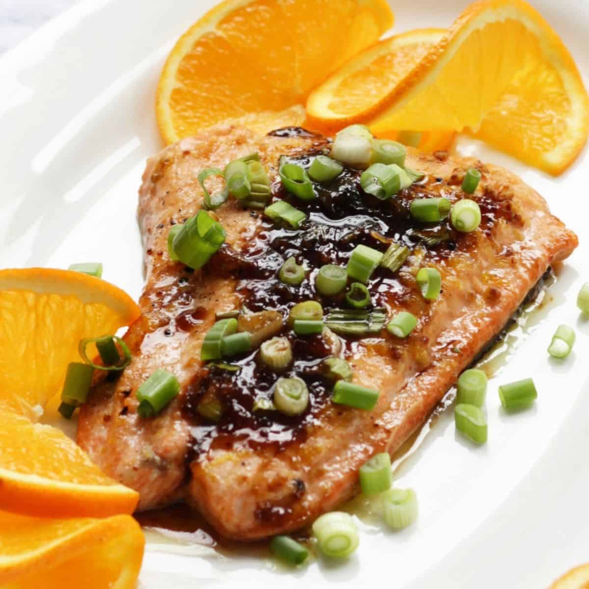 glistening salmon topped with brown sauce and chopped green onions, surrounded by thin orange slices, arranged on a white plate