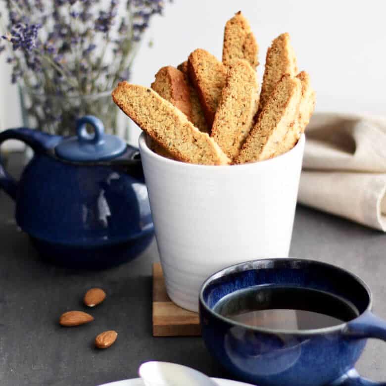biscotti standing upright in a white cup with a cup of tea in front and lavender behind