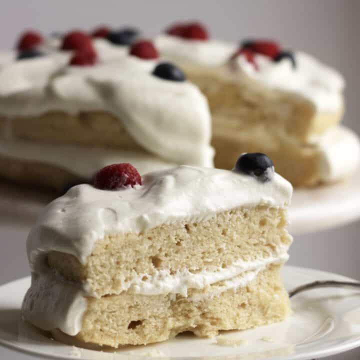 closeup of wedge of gluten free vanilla cake topped with whipped cream and berries, remaining cake in background