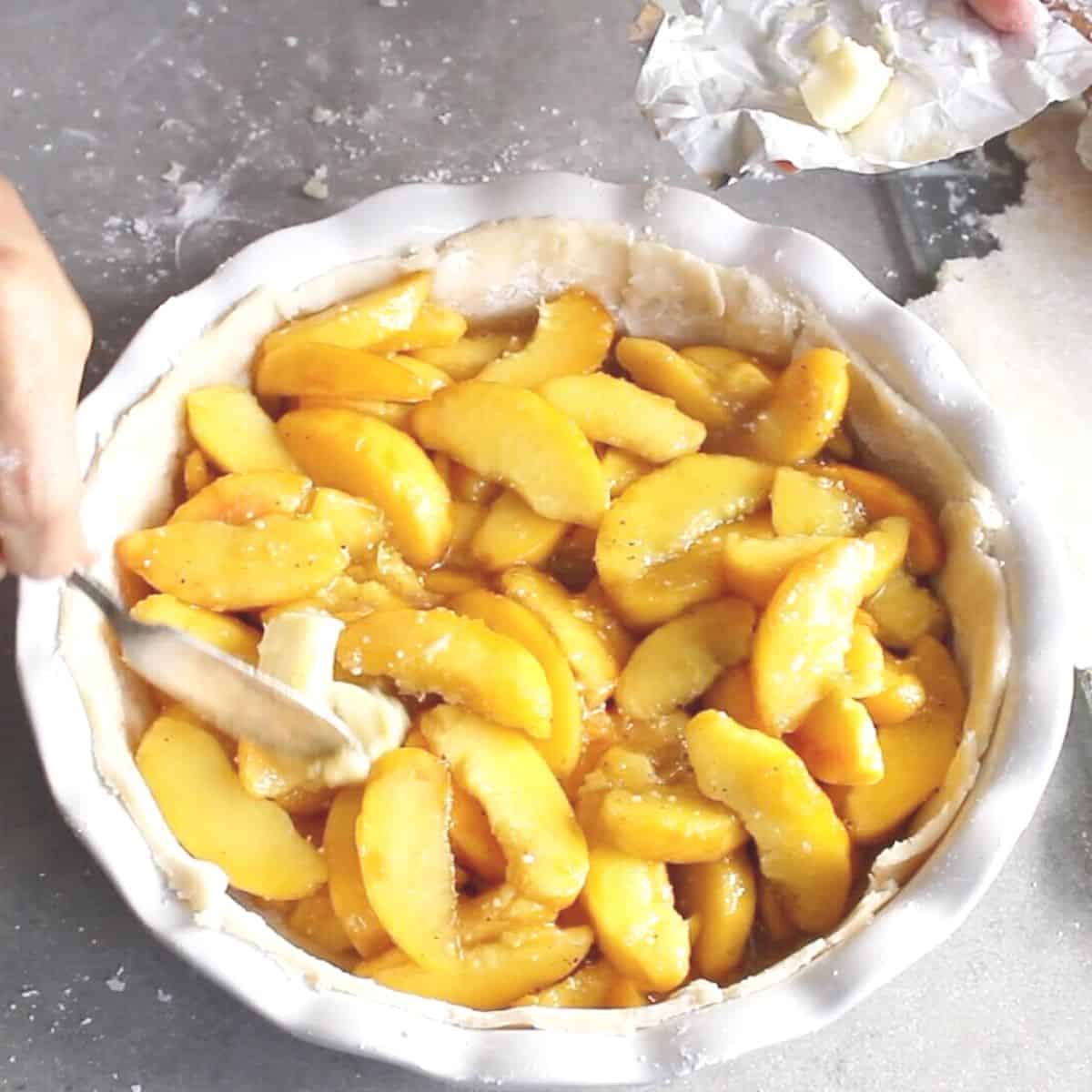 butter is dabbed onto peach slices in unbaked pie shell.