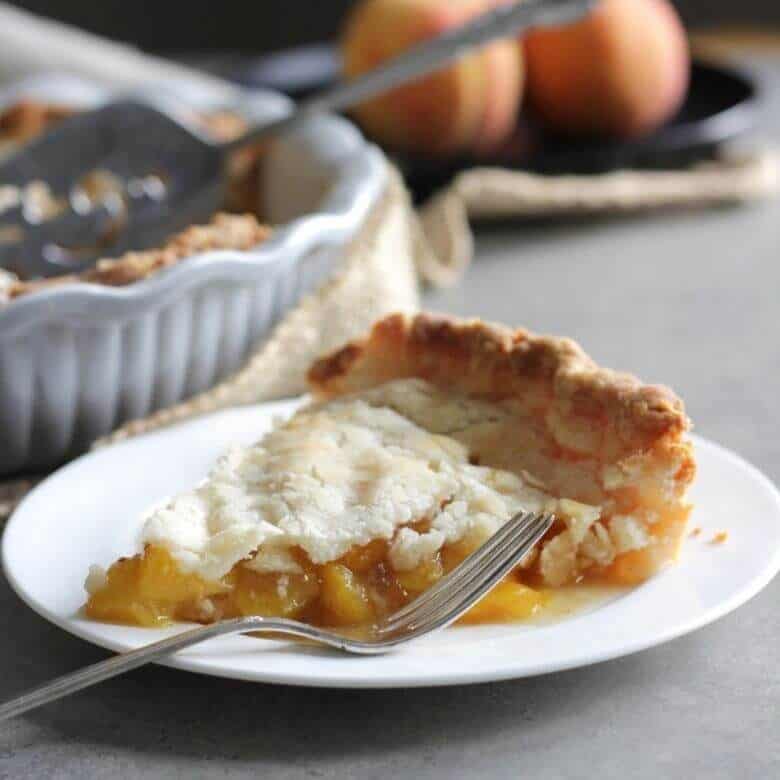 single slice of gluten free peach pie with whole pie and fresh peaches in the background