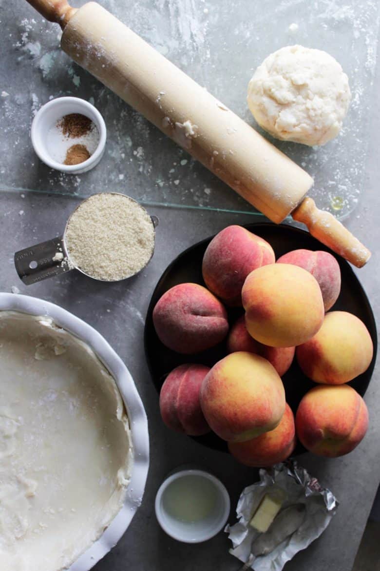 flat lay of fresh peaches, unbaked crust in pie plate, sugar, rolling pin, and other ingredients