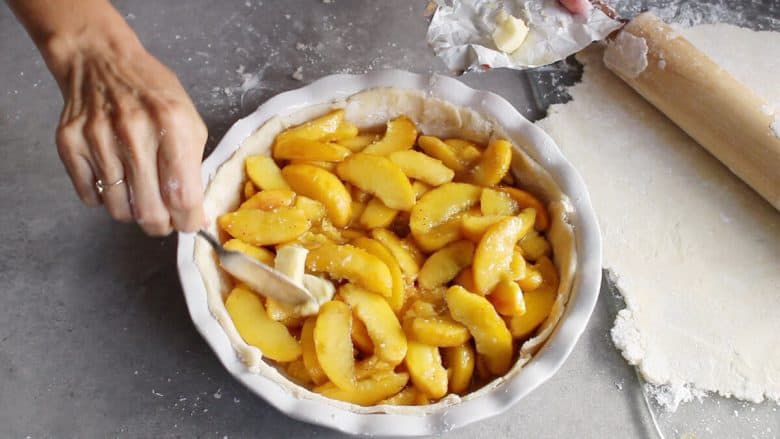butter is patted onto peach slices in unbaked pie crust
