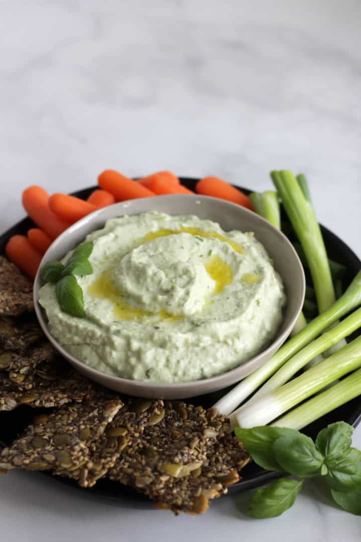 shallow bowl of cream, greenish avocado dip surrounded by seed crackers, baby carrots, and green onions