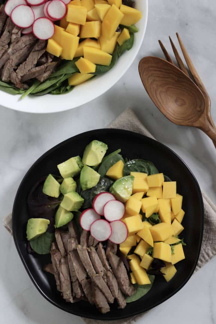 looking down on a black plate of beef salad with wooden spoon and more salad in background