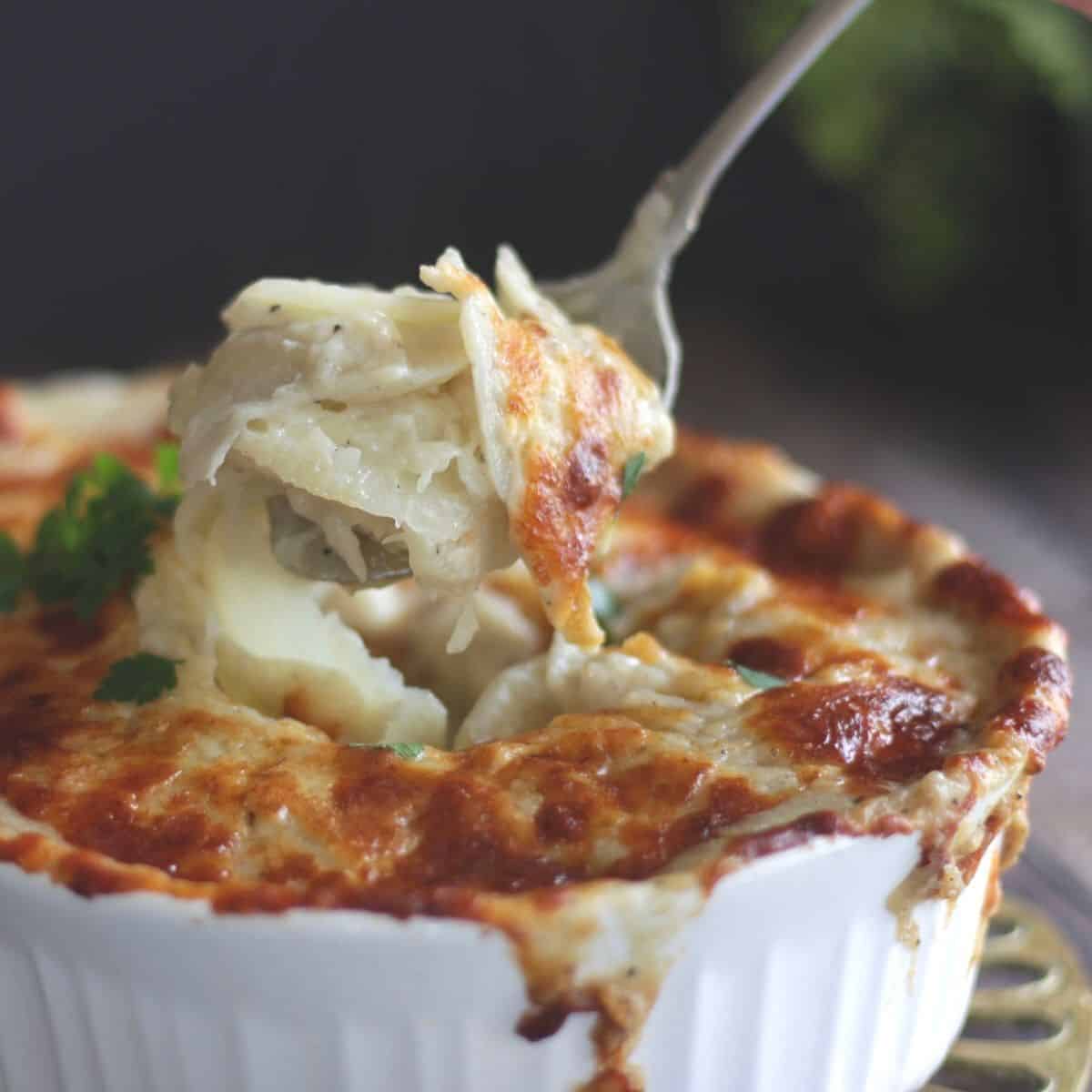 fork lifting sliced scalloped potatoes from white casserole dish dripping with melted cheese