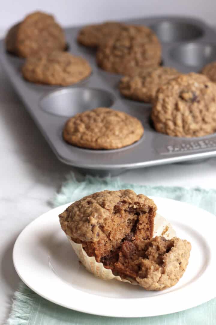 broken open applesauce muffin in parchment paper on white plate with remaining muffins in tin in background