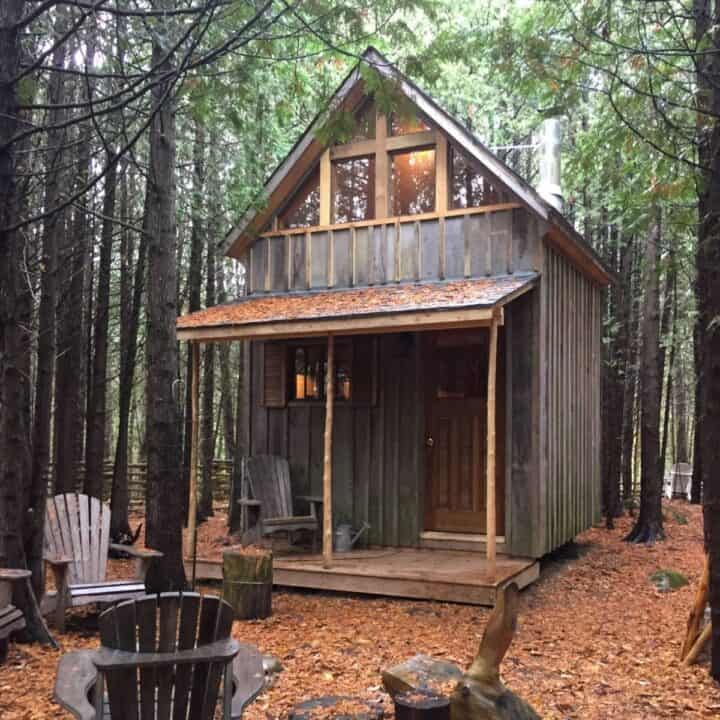 small cabin clad in grey wood, with porch on front, set in woods