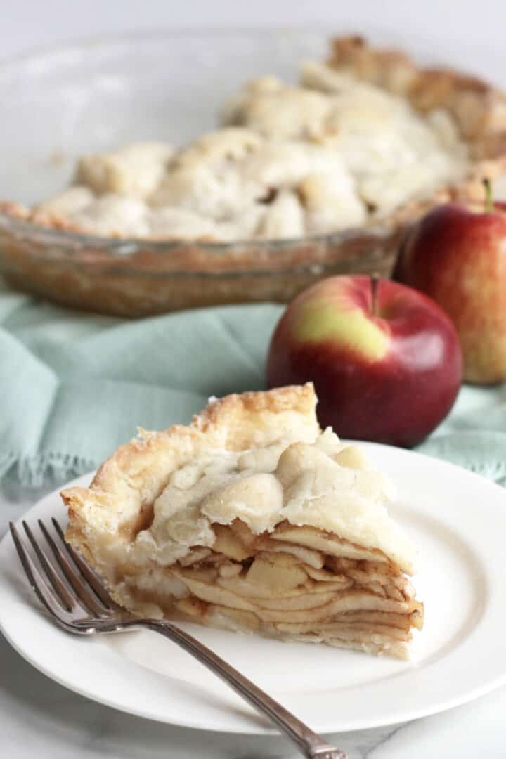 wedge of gluten free apple pie on white plate in front of apples and remaining pie