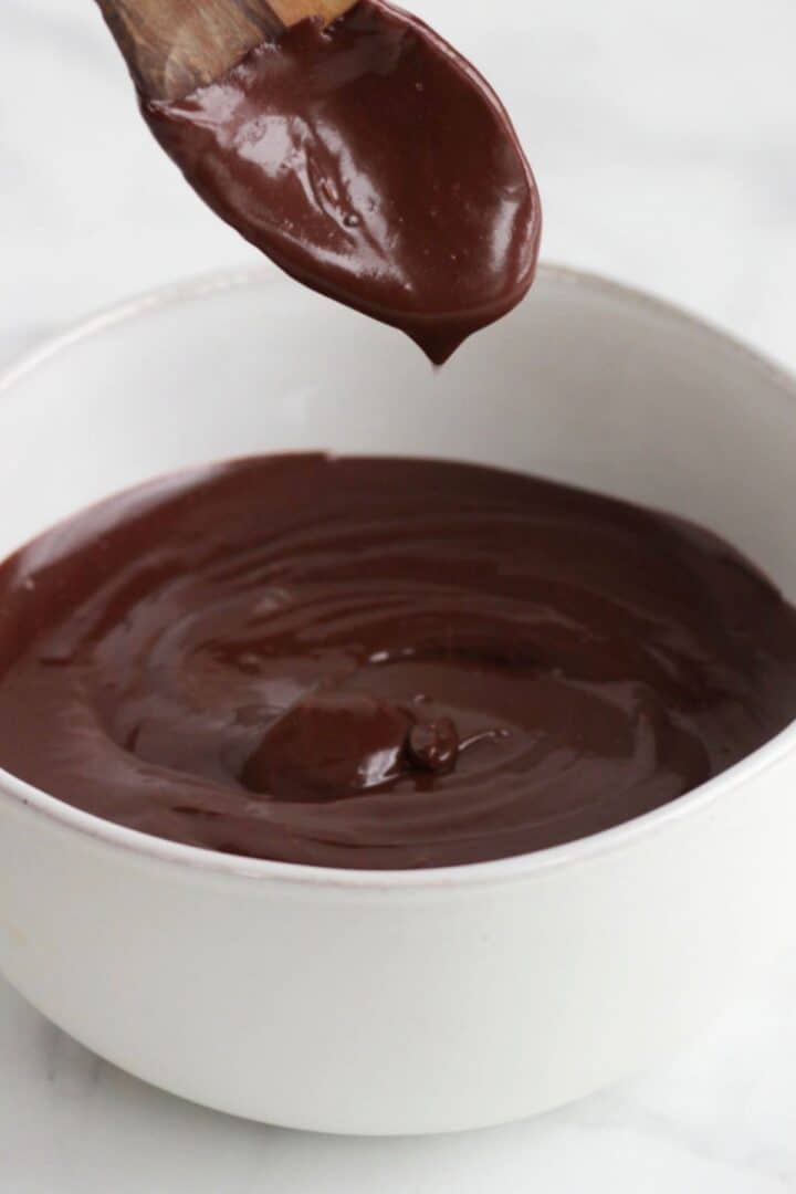 thick chocolate ganache coating a spoon held over a white bowl of the icing