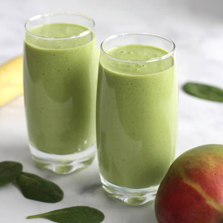 two glasses of green smoothies and partial mango, banana, and spinach leaves around them