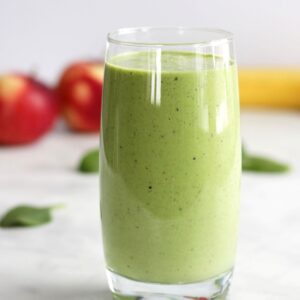 glass of green smoothie with apples and banana in background