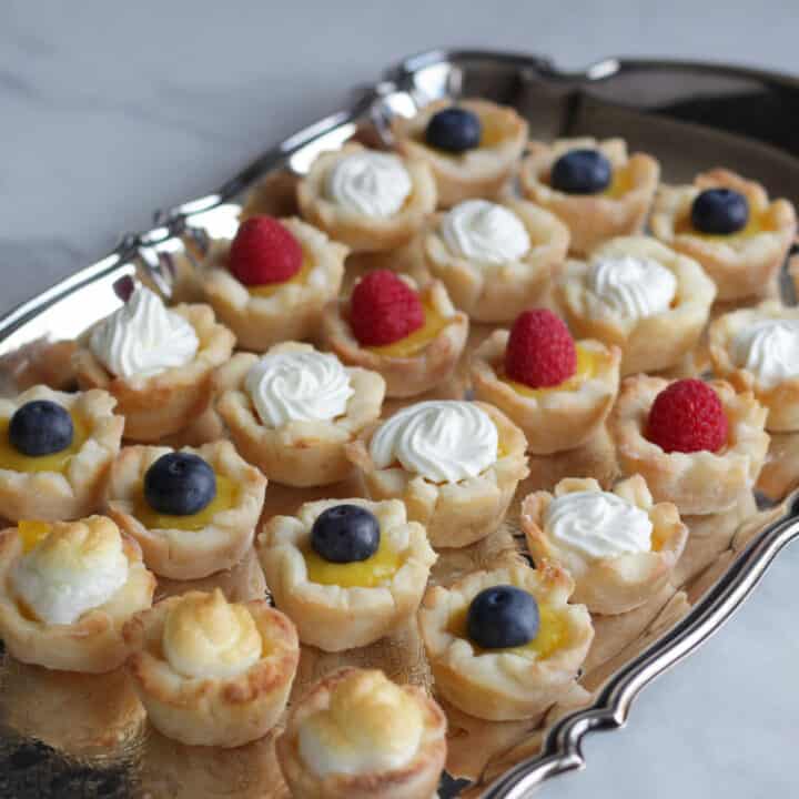 rows of mini lemon tartlets garnished with berries, whipped cream, and meringue on silver tray