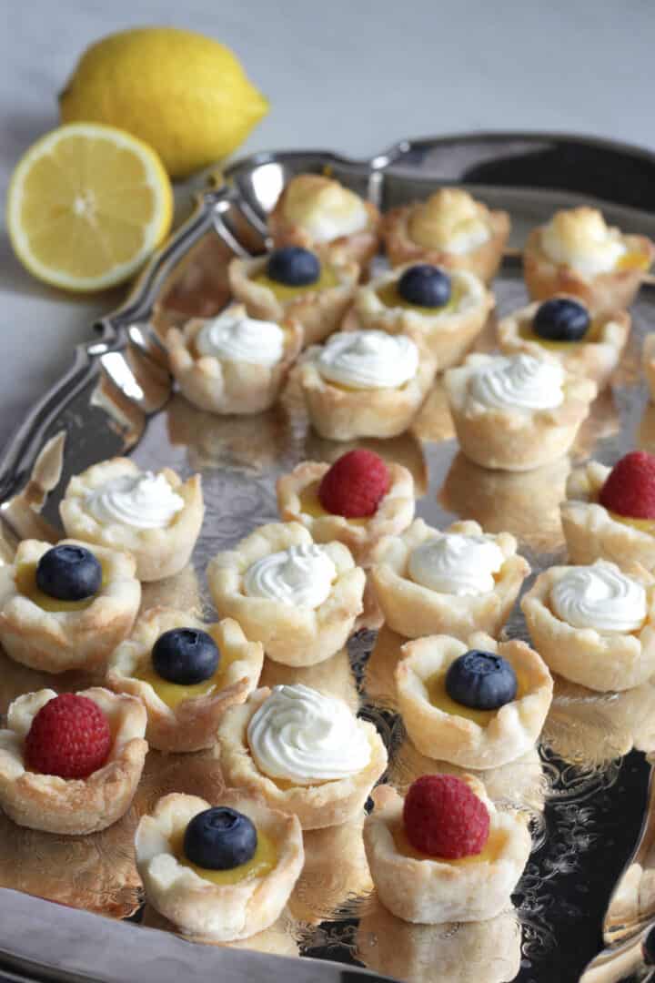 mini lemon tarts garnished with berries, whipped cream, and meringue sit on a silver tray with a couple fresh lemons in background