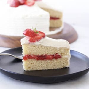 closeup of a slice of two-layer vanilla cake with strawberry filling, while frosting, and sliced fresh strawberries on top.