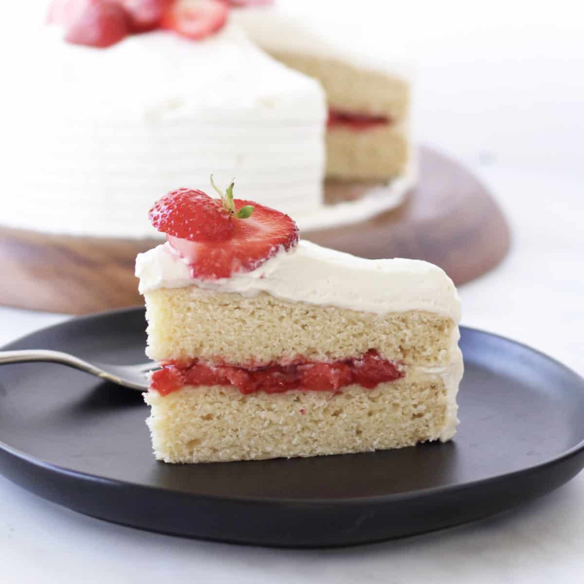 closeup of a slice of two-layer vanilla cake with strawberry filling, while frosting, and sliced fresh strawberries on top.