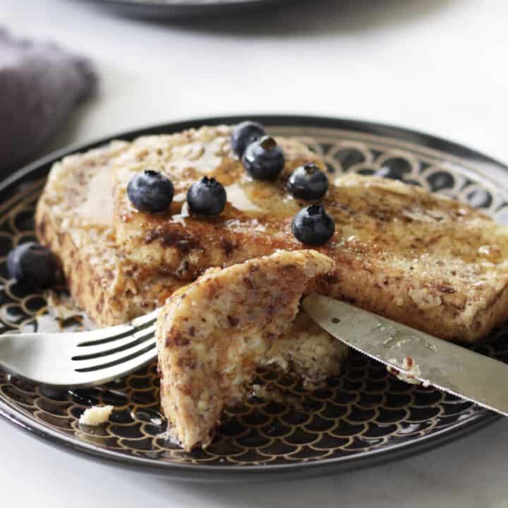 knife and fork taking a bite of golden brown French toast with blueberries and maple syrup on it