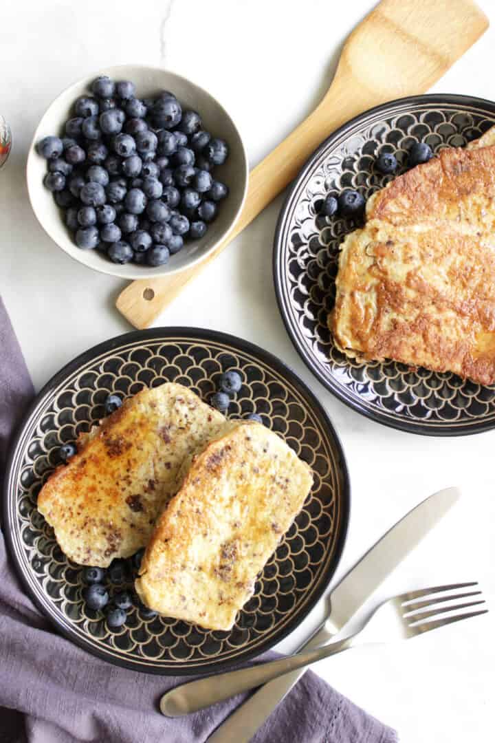 golden French toast on black plates by a bowl of blueberries, seen from above