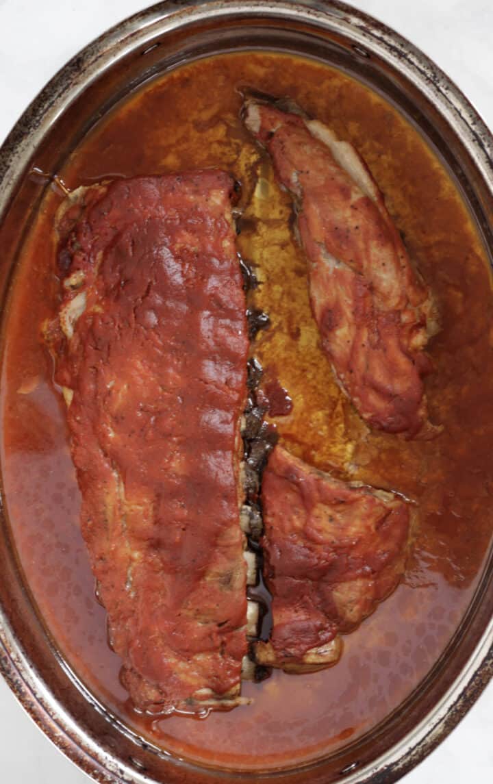 baked rack of ribs sitting in a pool of red sauce in bottom of roasting pan