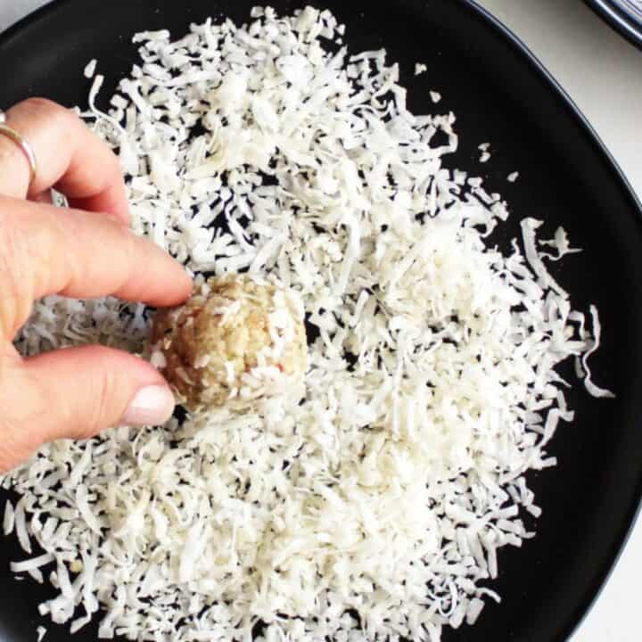 coconut ball is rolled by fingers in flaked coconut scattered on black plate