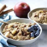 two bowls of apple cinnamon quinoa porridge, garnished with blueberries, whole apple and cinnamon sticks in background