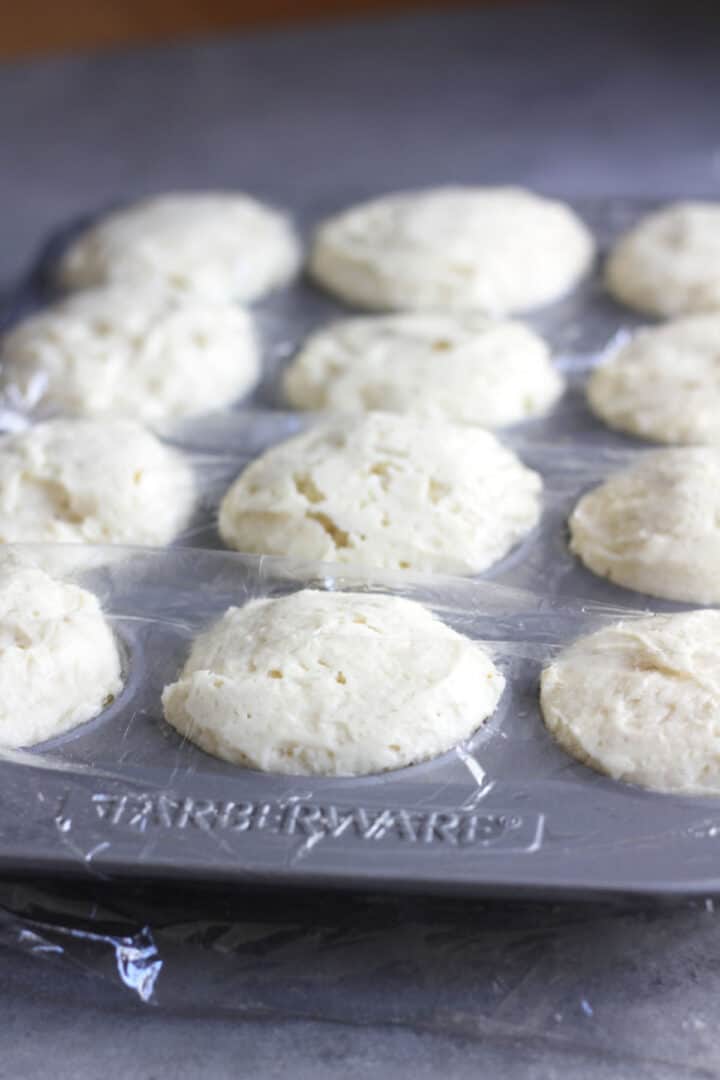 risen white dough in muffin pan, covered in plastic wrap