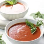 two white bowls of shimmering red soup, garnished with fresh basil