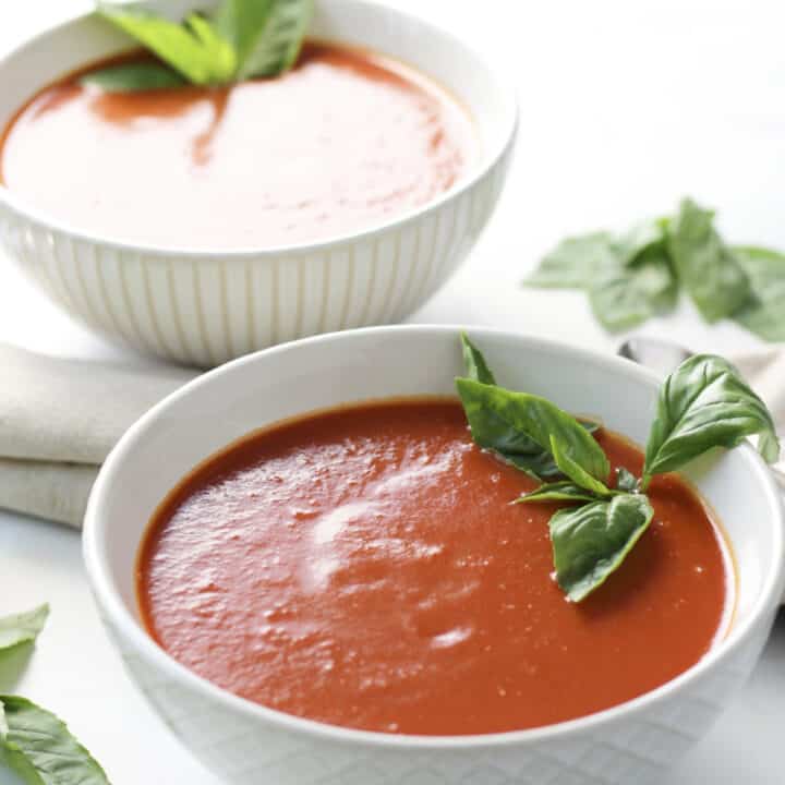 two white bowls of shimmering red soup, garnished with fresh basil