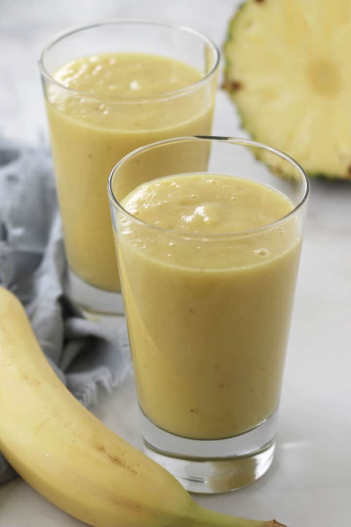 two glasses of creamy yellow smoothie with part of a banana and a cut pineapple around them