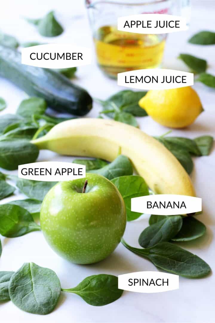 apple juice in pyrex measuring cup, English cucumber, lemon, banana, green apple, and spinach leaves scattered on white surface