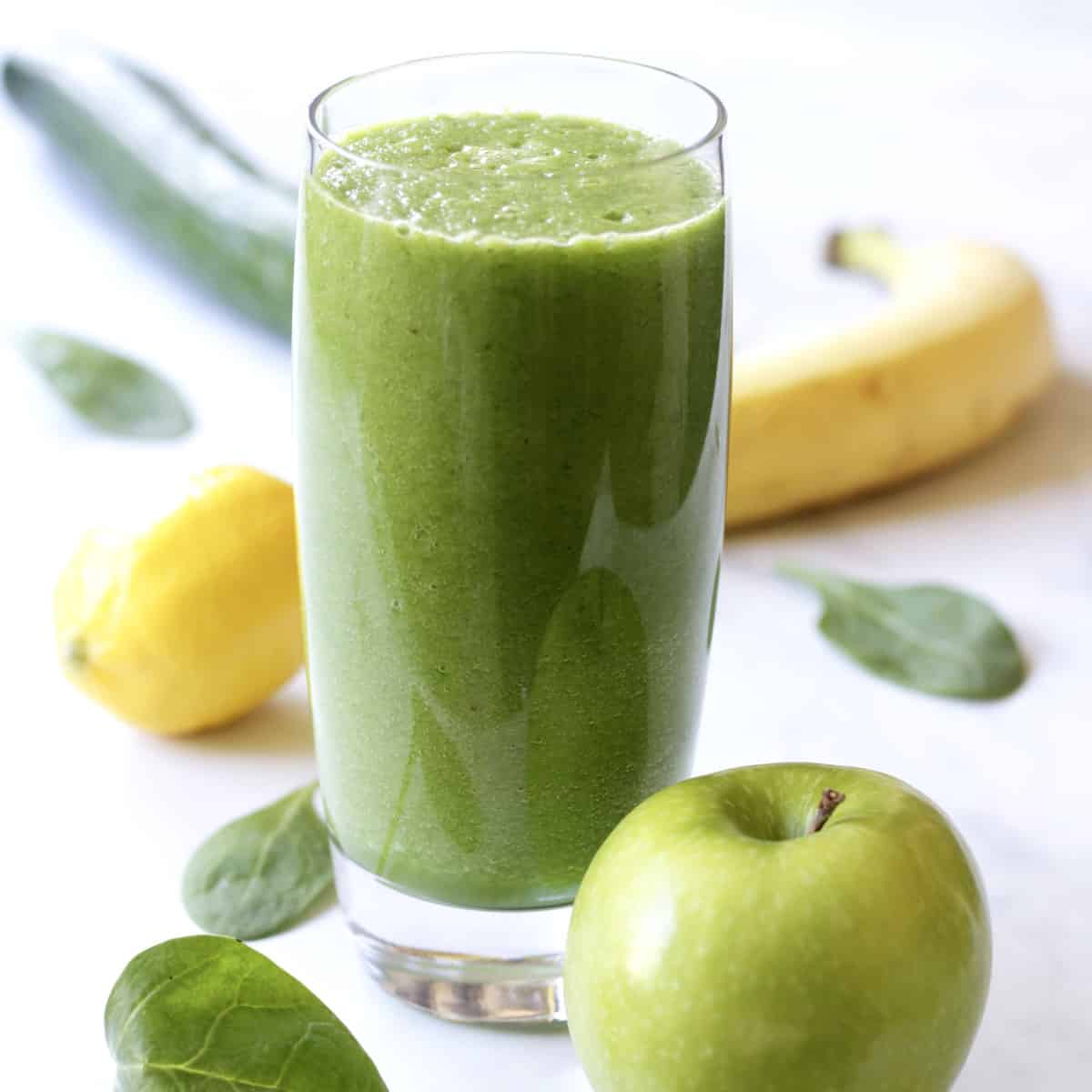 green smoothie in glass surrounded by green apple, banana, lemon, cucumber, and spinach leaves