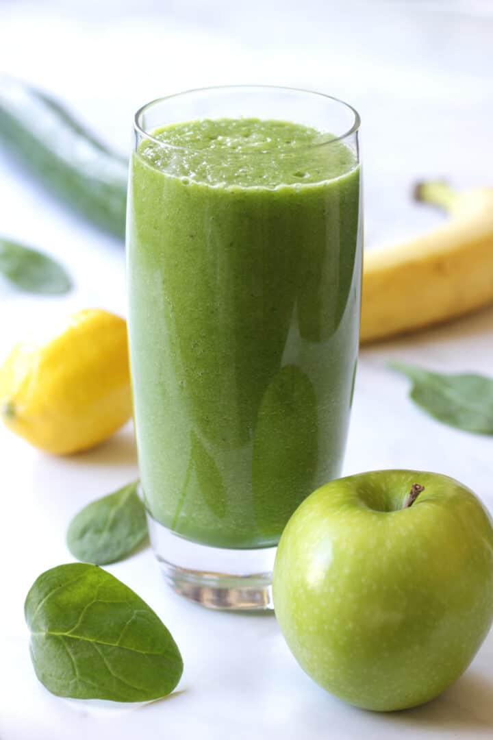 green smoothie in glass with green apple, banana, lemon, cucumber, and spinach leaves around it