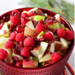 chopped green apples, pears, grapes, and kiwi with red apples, pear, grapes, and raspberries in festive red bowl with greenery in background