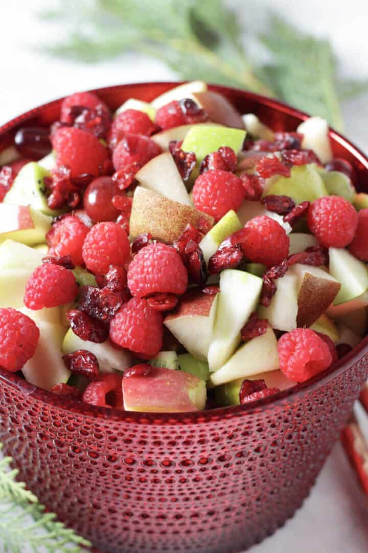 glass bowl filled with chopped fruit and berries, surrounded by greenery