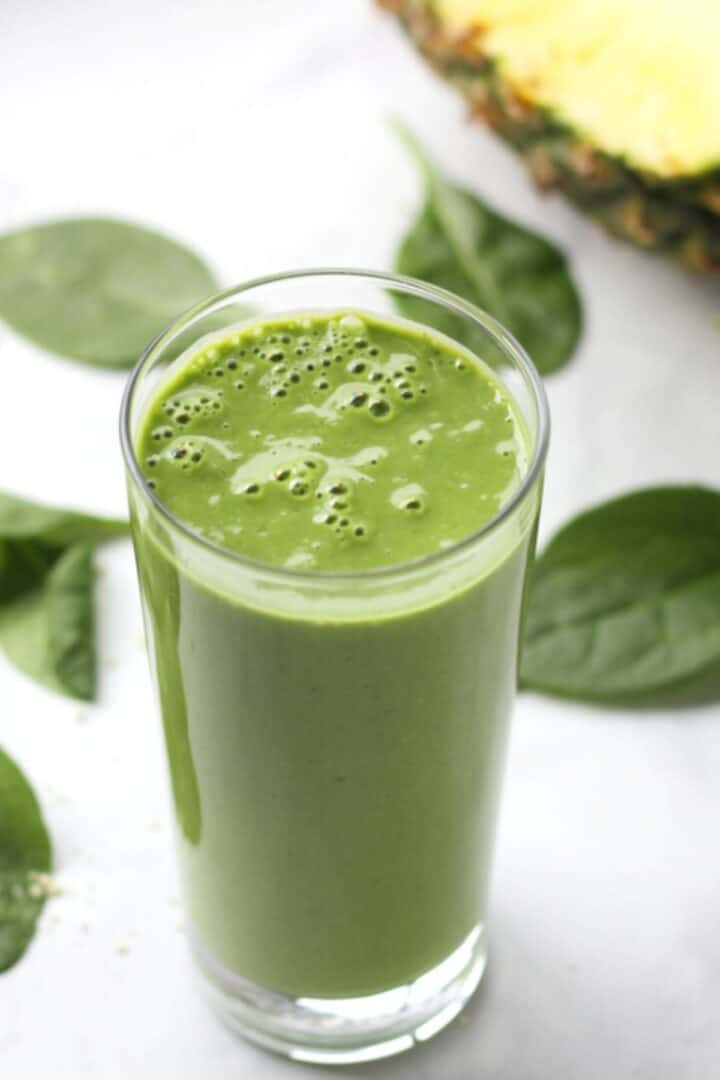 tall glass of green smoothie with partially-cut pineapple and loose spinach leaves around it