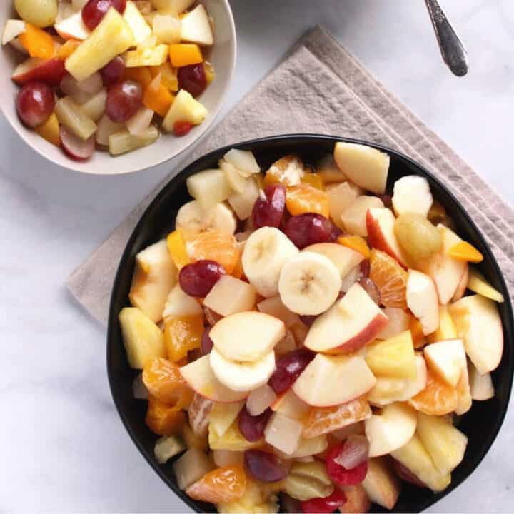 top-down view of a black bowl filled with chunks of apples, bananas, oranges, pineapple, and grapes