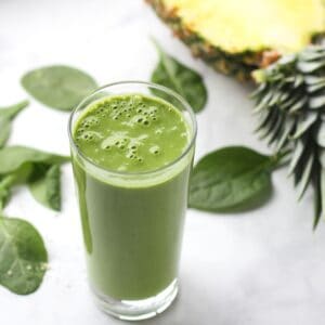 glass filled with a fresh green smoothie with bubbles breaking the surface, spinach leaves, and pineapple in background