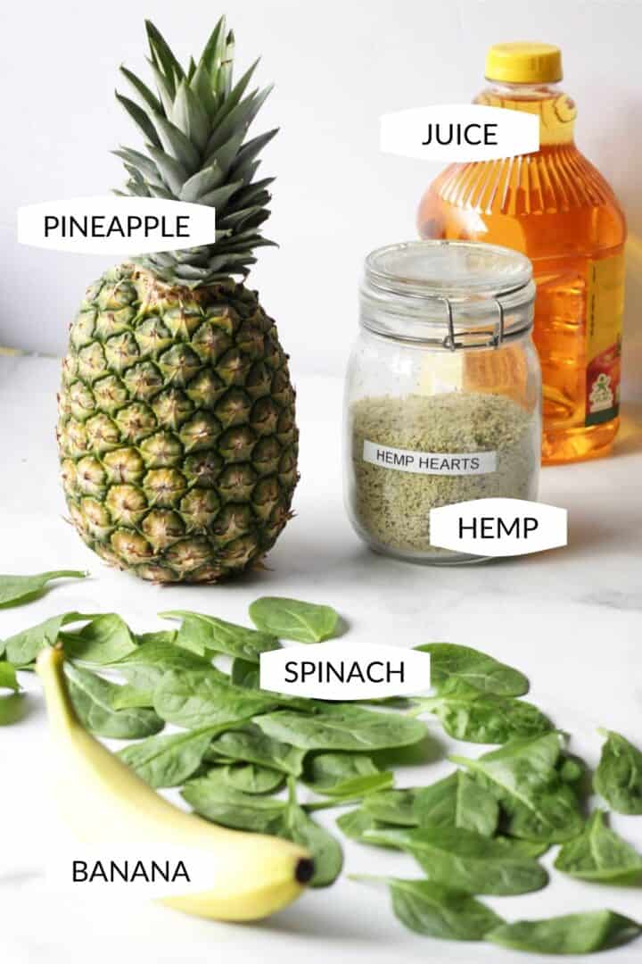 whole, fresh pineapple, jar of hemp seeds, bottle of apple juice, loose spinach leaves, and a banana on white marble surface
