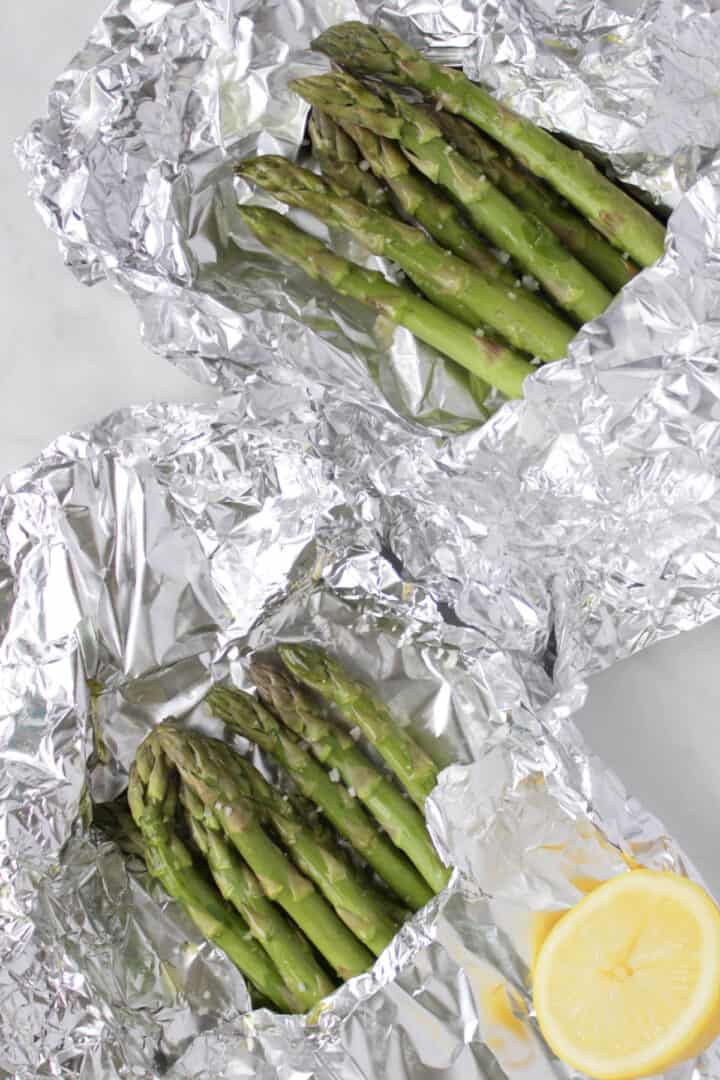 two open packets of foil-wrapped asparagus with a cut open lemon.