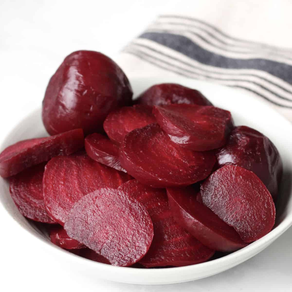 cooked sliced beets and a whole one in a shallow white bowl with a neutral striped tea towel in background